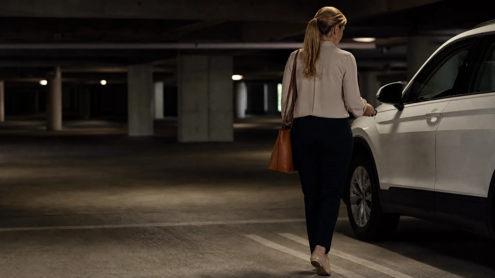A woman in a light blouse and dark trousers with a brown shoulder bag walks toward a white SUV in a dimly lit concrete parking garage.