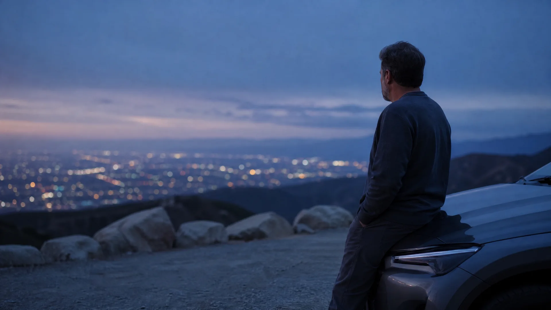 A man in a dark long-sleeve shirt leans against the front of a light-colored SUV at a gravel overlook at dusk, facing a valley filled with city lights.